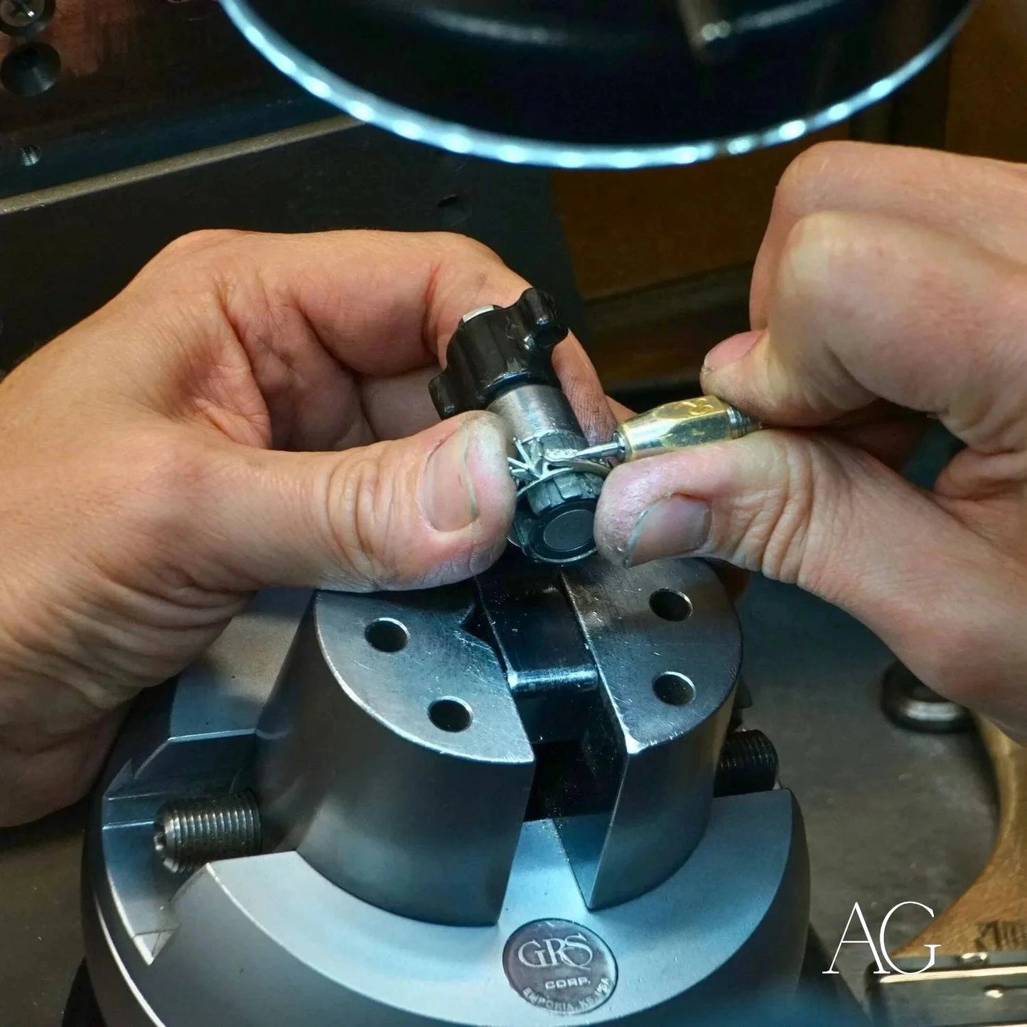 Hands repairing gold jewelry with a small mechanical tool on a circular metal base