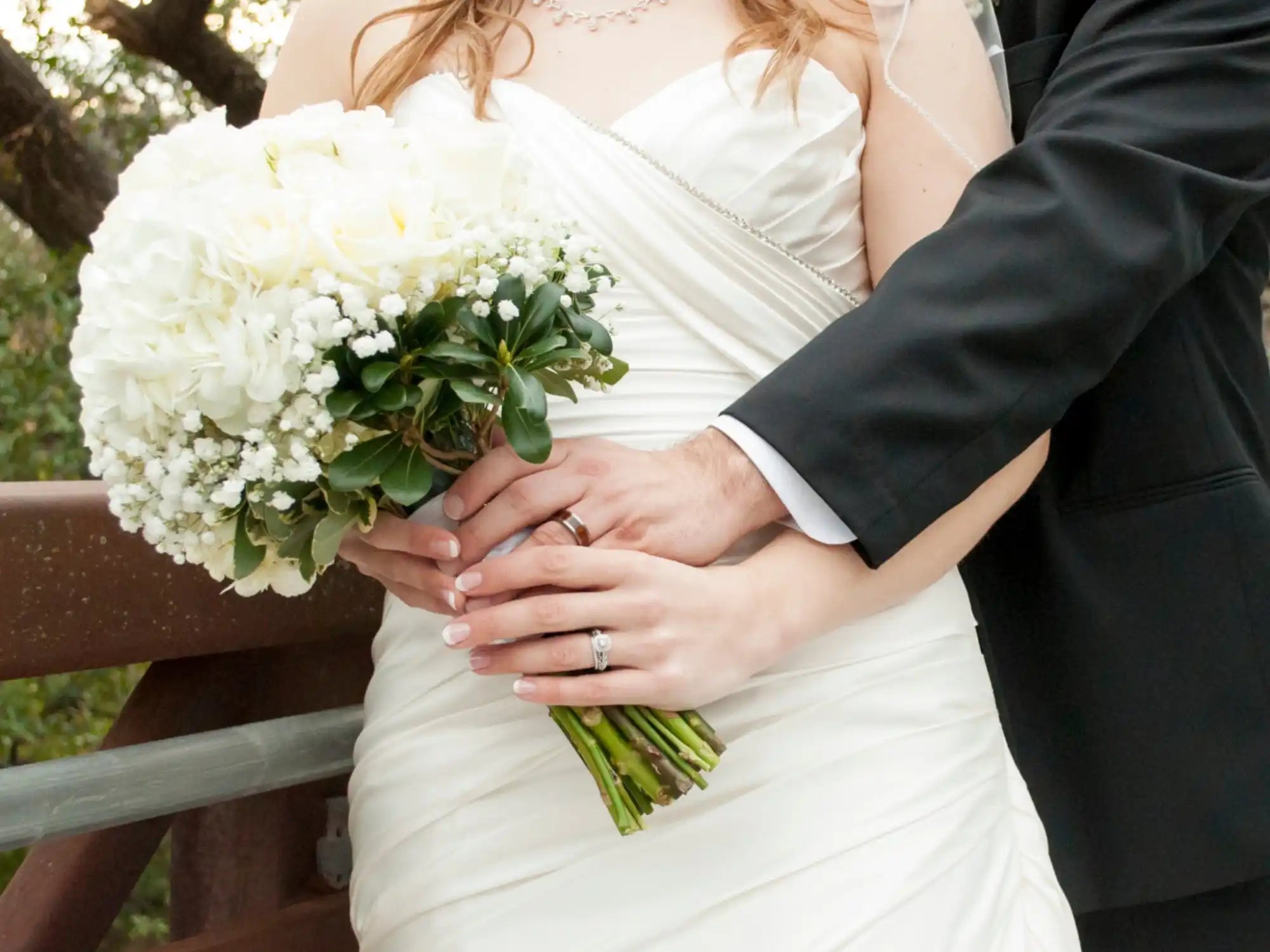 Beautiful white wedding bouquet with baby’s breath and greenery held by hands wearing wedding rings.