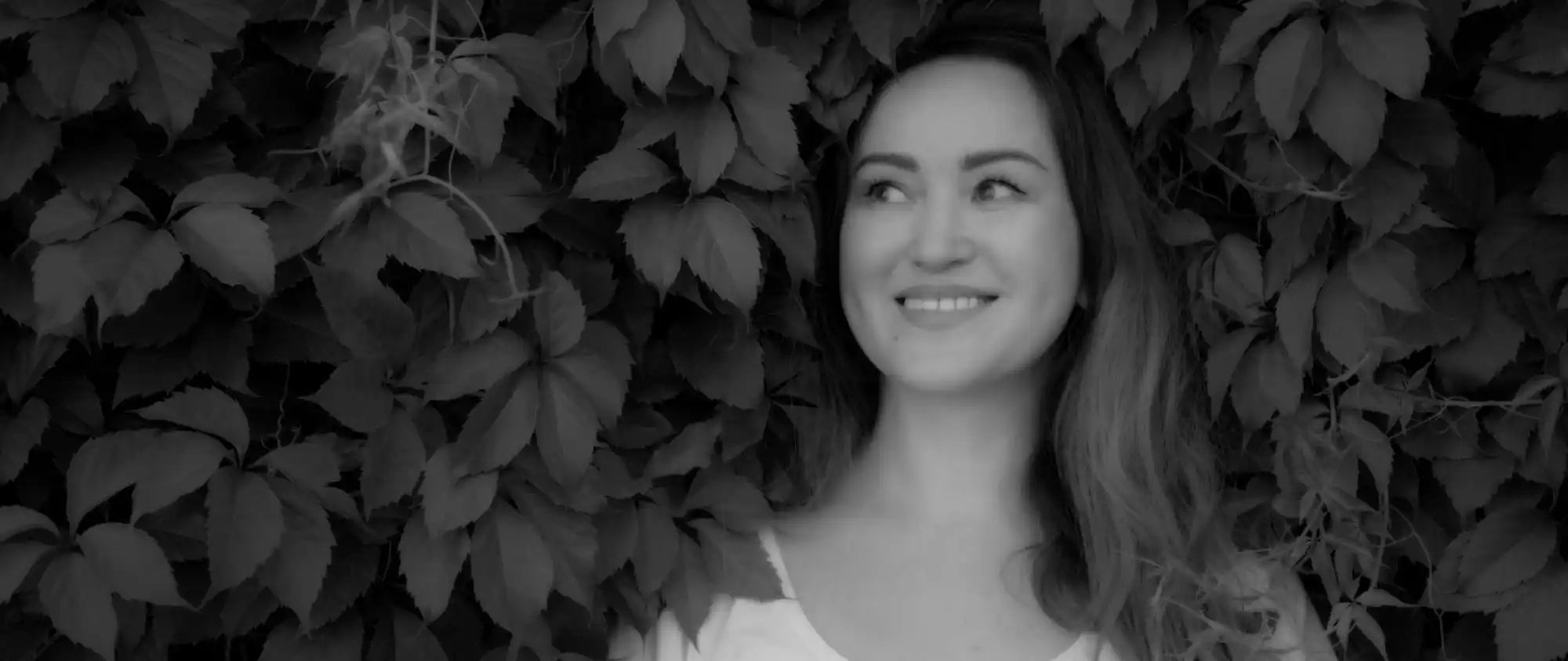 A black and white portrait of someone smiling with long hair against foliage.