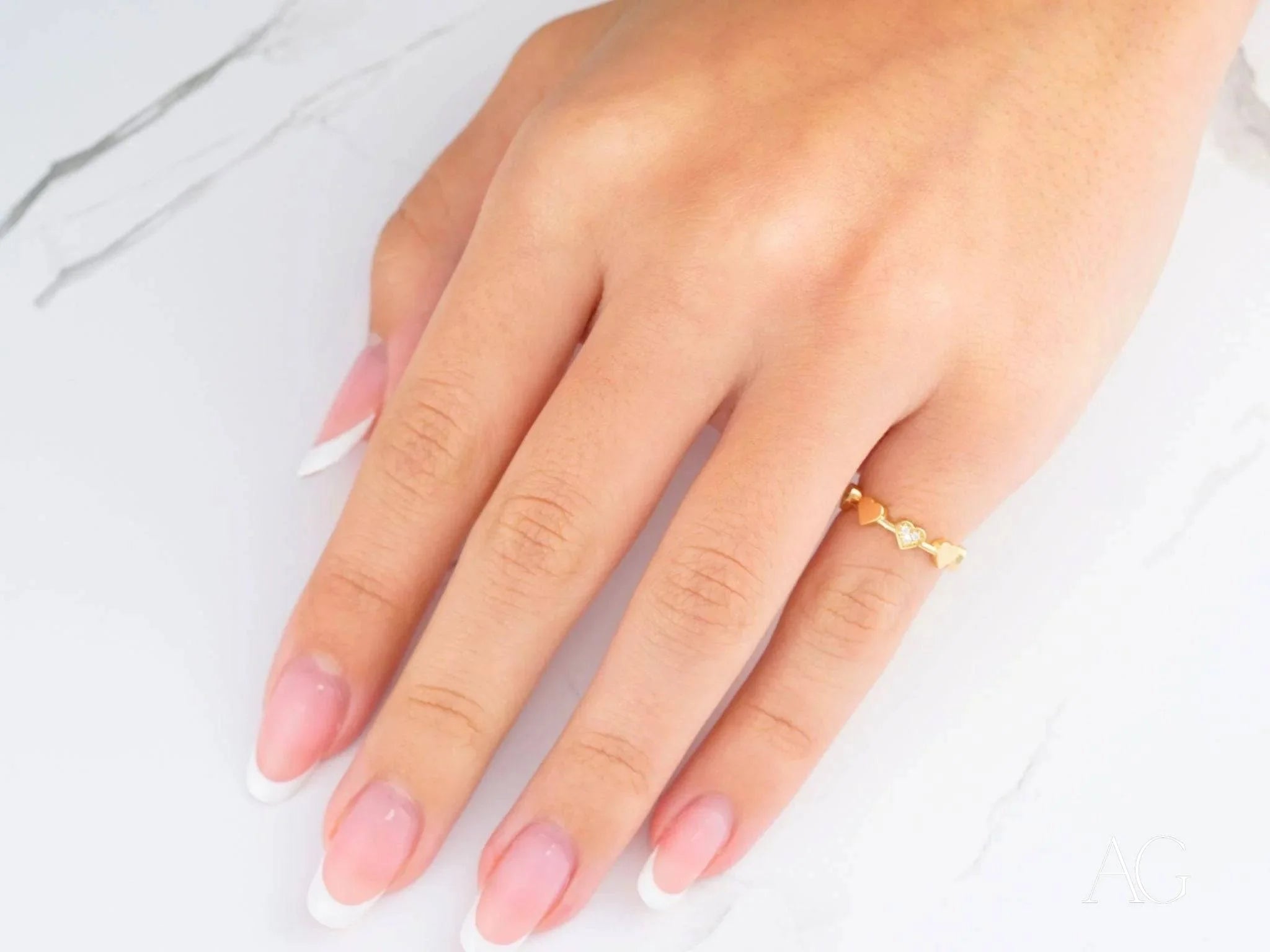 Hand showcasing a delicate 18k yellow gold CZ heart ring with pink manicured nails