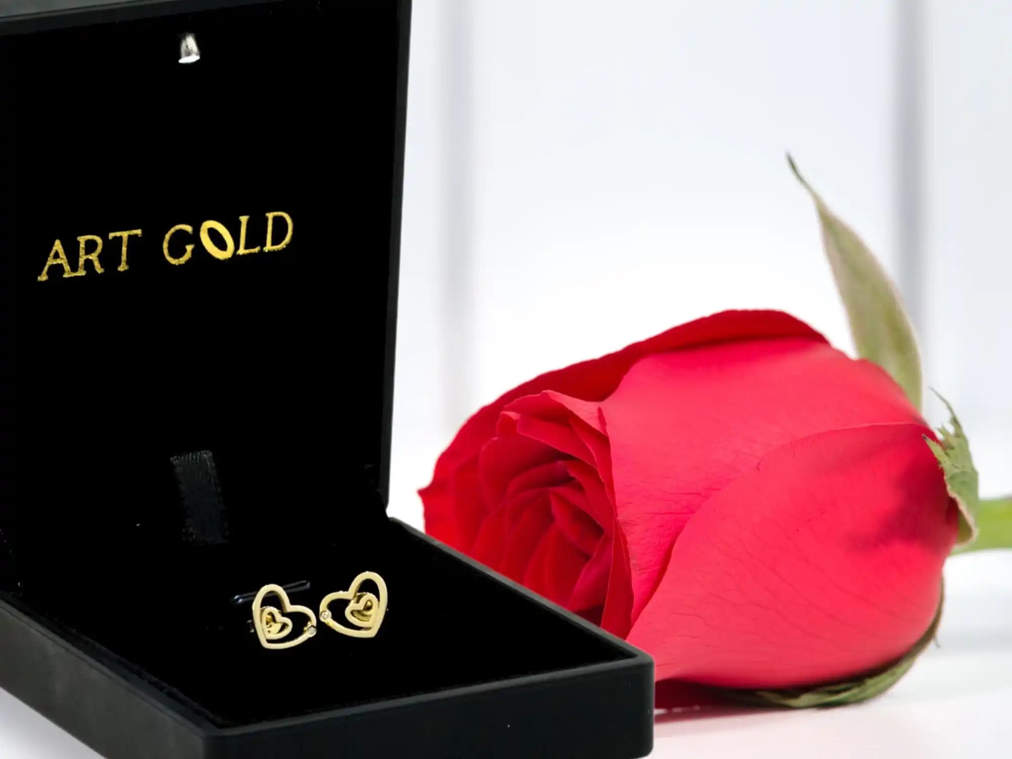 Gold heart-shaped earrings in a black jewelry box alongside a red rose.