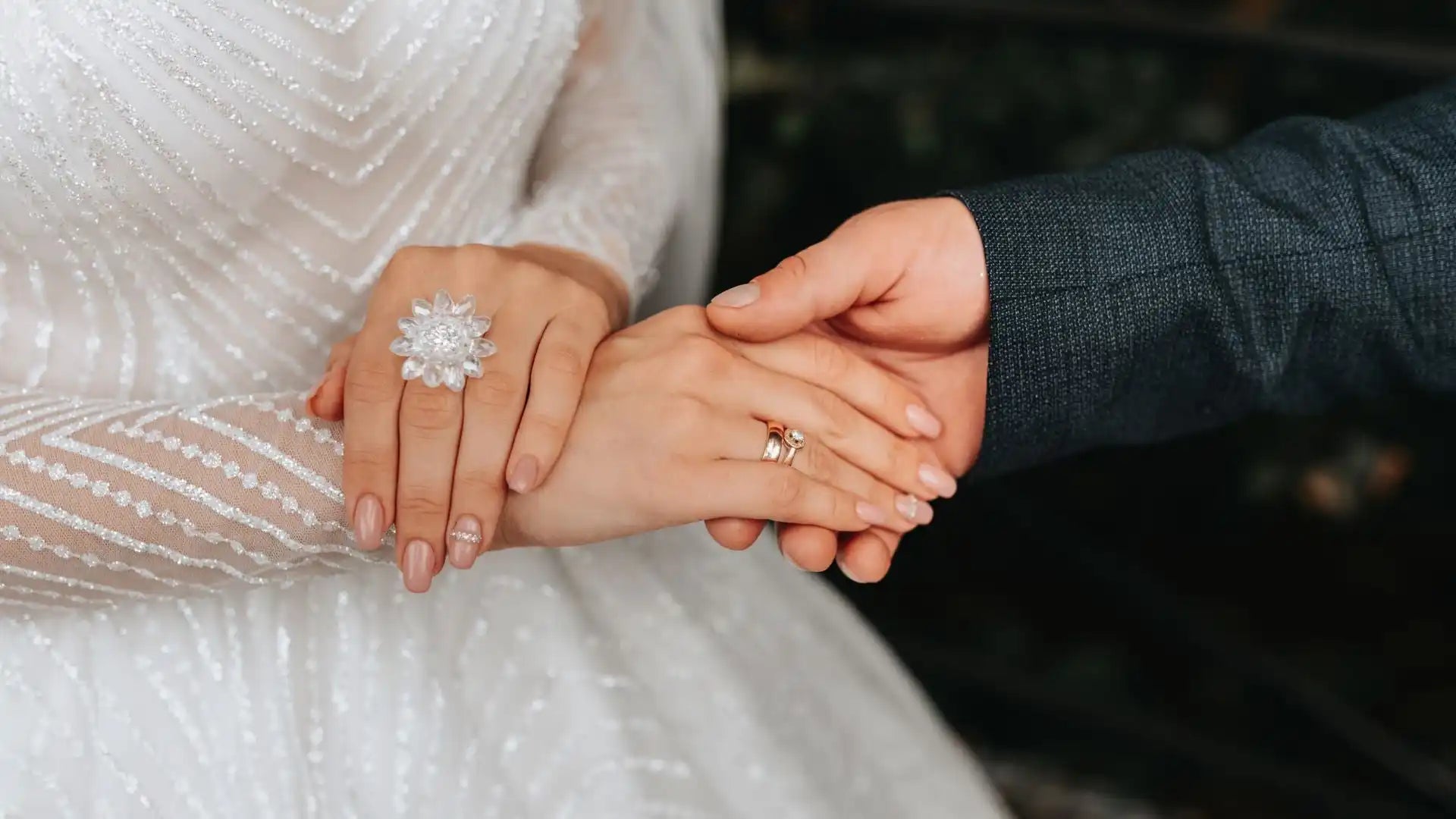 Two hands clasped together, with one wearing a white lace sleeve and a statement ring while the other wears a dark suit sleeve.